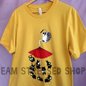 May include: Mustard yellow t-shirt featuring a whimsical illustration. A teapot pours liquid onto a red umbrella, with black cartoon characters below. The shirt has a crew neck and short sleeves. The text "SEAM STRESSED SHOP" is printed at the bottom.
