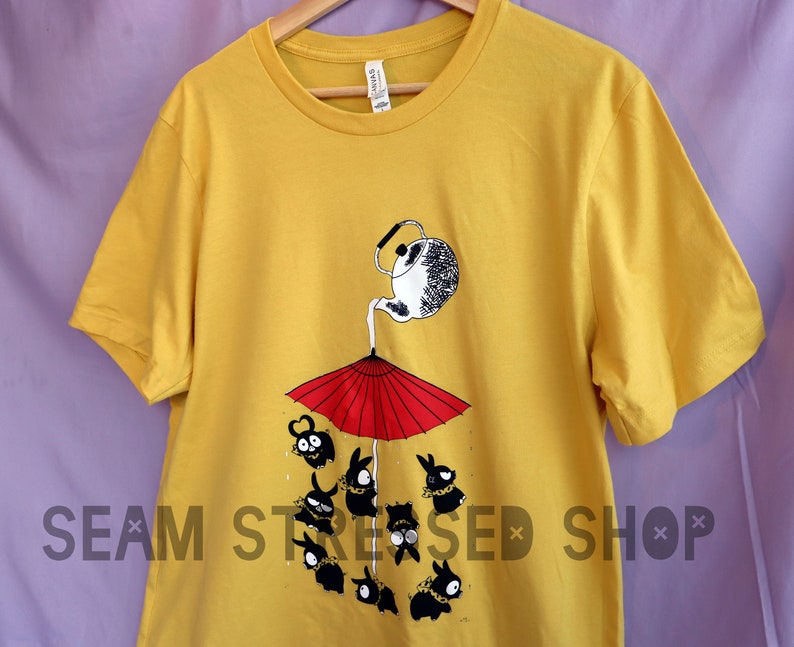 May include: Mustard yellow t-shirt featuring a whimsical illustration. A teapot pours liquid onto a red umbrella, with black cartoon characters below. The shirt has a crew neck and short sleeves. The text "SEAM STRESSED SHOP" is printed at the bottom.