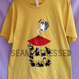 May include: Mustard yellow t-shirt featuring a whimsical illustration. A teapot pours onto a red umbrella, sheltering several black bunny-like creatures. The shirt has a crew neck and short sleeves. The brand tag is visible.