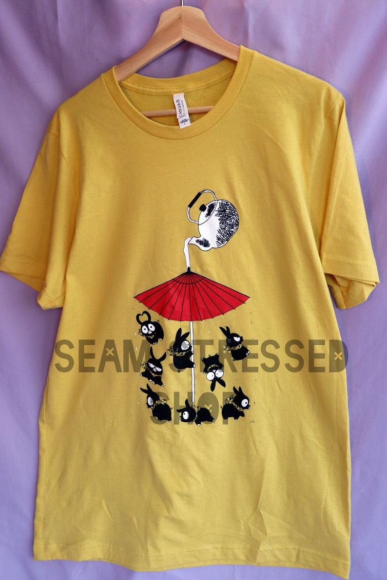 May include: Mustard yellow t-shirt featuring a whimsical illustration. A teapot pours onto a red umbrella, sheltering several black bunny-like creatures. The shirt has a crew neck and short sleeves. The brand tag is visible.