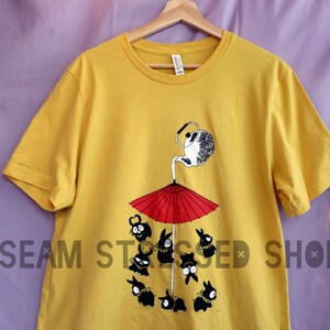 May include: Mustard yellow t-shirt featuring a whimsical illustration. A teapot pours liquid onto a red umbrella, with cartoon figures below. The text "SEAM STRESSED SHOP" is printed on the shirt.