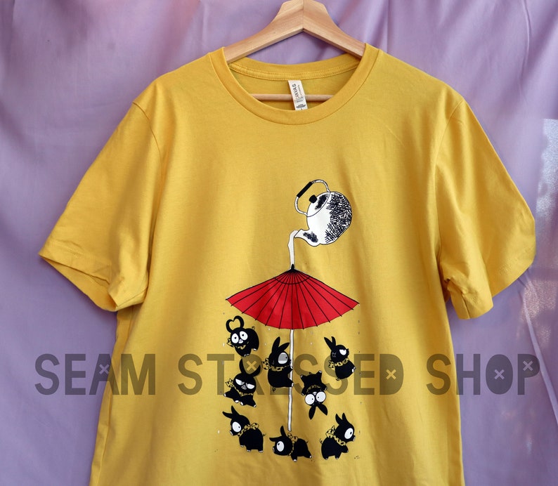 May include: Mustard yellow t-shirt featuring a whimsical illustration. A teapot pours liquid onto a red umbrella, with cartoon figures below. The text "SEAM STRESSED SHOP" is printed on the shirt.