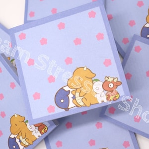May include: A stack of light blue square sticky notes with a light purple border. Each note features a cartoon illustration of a yellow creature with a white and red creature. Pink flower shapes are scattered across the surface.
