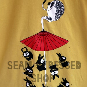 May include: Mustard yellow t-shirt featuring a whimsical illustration. A white teapot pours onto a red umbrella, sheltering several black rabbit-like creatures. The creatures wear scarves and have various expressions.