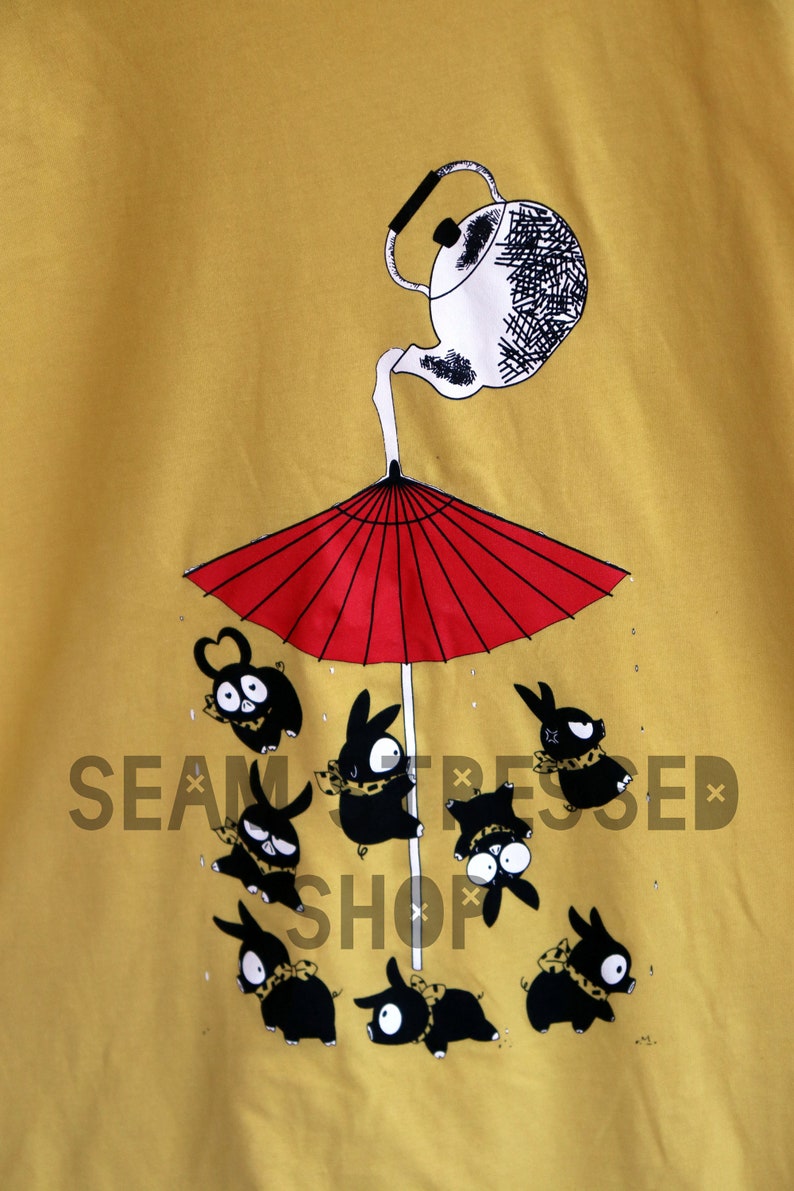 May include: Mustard yellow t-shirt featuring a whimsical illustration. A white teapot pours onto a red umbrella, sheltering several black rabbit-like creatures. The creatures wear scarves and have various expressions.