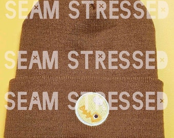 May include: Brown knit beanie with a circular patch featuring a yellow and orange design. The text "SEAM STRESSED" is overlaid on the beanie. The beanie is displayed against a yellow background.