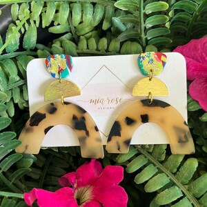 May include: A pair of statement earrings with a colorful abstract top, a gold-colored half-circle, and a tortoise-shell patterned arch. The earrings are displayed on a white card with the text "mia rose designs".