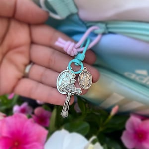 May include: A silver charm with a cross, a guardian angel, and the Virgin Mary. The charm is attached to a blue and pink zipper pull.