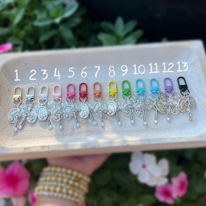 May include: Thirteen silver keychains with a cross and a religious icon. The keychains are numbered 1 through 13 and have different colored clasps: white, pink, red, orange, yellow, green, blue, light blue, dark blue, black.