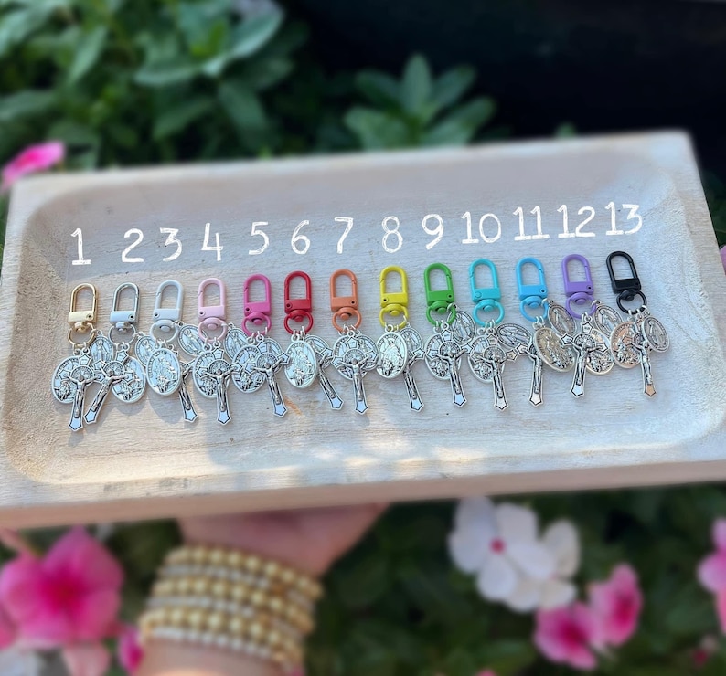 May include: Thirteen silver keychains with a cross and a religious icon. The keychains are numbered 1 through 13 and have different colored clasps: white, pink, red, orange, yellow, green, blue, light blue, dark blue, black.