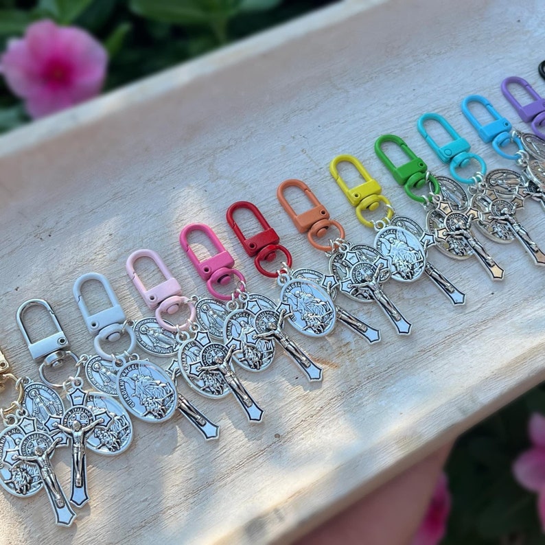 May include: A set of 10 colorful keychains with silver charms featuring a religious image of a cross and a saint. The keychains are arranged in a row on a wooden surface.