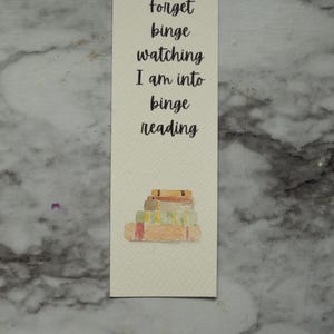 May include: A rectangular bookmark with the text "Forget binge watching I am into binge reading" in black script. A watercolor illustration of a stack of books is at the bottom. The bookmark is on a marble surface.