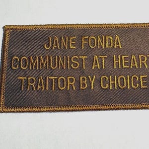 May include: A rectangular patch with the embroidered text "JANE FONDA COMMUNIST AT HEART TRAITOR BY CHOICE" in gold thread on a brown background. The patch has a gold border.