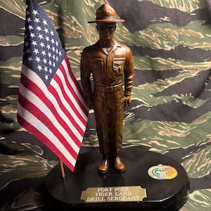 May include: A brown statue of a soldier in uniform, wearing a hat, standing on a black base. An American flag is to the left. The base has a gold plaque that reads "FORT POLK TIGER LAND DRILL SERGEANT".