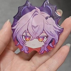 May include: A glossy acrylic pin featuring a stylized anime-inspired character head with light purple hair, pointed dark purple horns, and vibrant orange and yellow eyes. The character has a small, neutral expression and is framed by a dark purple border.