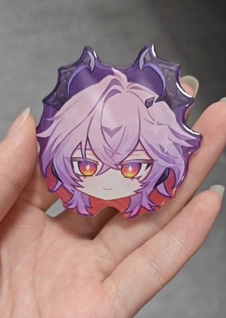 May include: A glossy acrylic pin featuring a stylized anime-inspired character head with light purple hair, pointed dark purple horns, and vibrant orange and yellow eyes. The character has a small, neutral expression and is framed by a dark purple border.