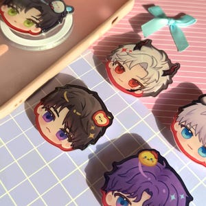 May include: A collection of colorful anime-style character phone grips. Each grip features a unique character design with vibrant hair and eyes. The grips are in various colors, including purple, black, and white, with cute accessories.