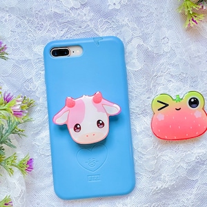 May include: A light blue smartphone case with a heart-shaped BT21 logo, featuring a pink and white cow-shaped phone grip attached to the center. Beside the case is a separate, winking frog-shaped phone grip with a red strawberry-like pattern.