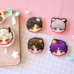 May include: A clear phone case with a pop socket featuring a cartoon character with white hair and red eyes. Four additional cartoon character face stickers are displayed on a pink and white striped surface.