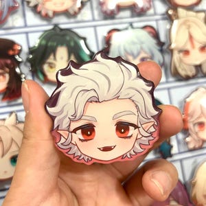 May include: A close-up of a white and red anime character pin. The character has white hair, red eyes, and a small, sharp, red fang. The pin is glossy and has a black outline.