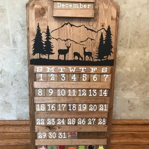May include: Wooden calendar with a mountain and deer design. The calendar is for December and has a white background with black numbers. The calendar also has a small section at the bottom with a variety of colorful icons.