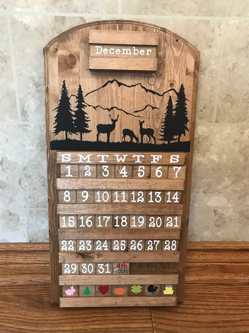 May include: Wooden calendar with a mountain and deer design. The calendar is for December and has a white background with black numbers. The calendar also has a small section at the bottom with a variety of colorful icons.