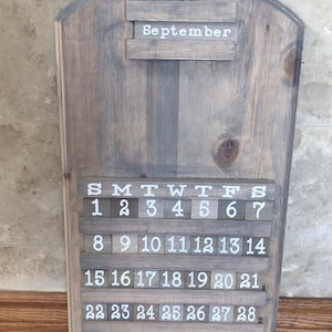 May include: A wooden calendar with a gray wash finish. The calendar features a white inscription for the month of September and a sliding block system for the days of the month. The calendar has a decorative row of icons at the bottom, including a shamrock, a pig, a birthday cake, a pumpkin, a turkey, a Christmas tree, and a heart.