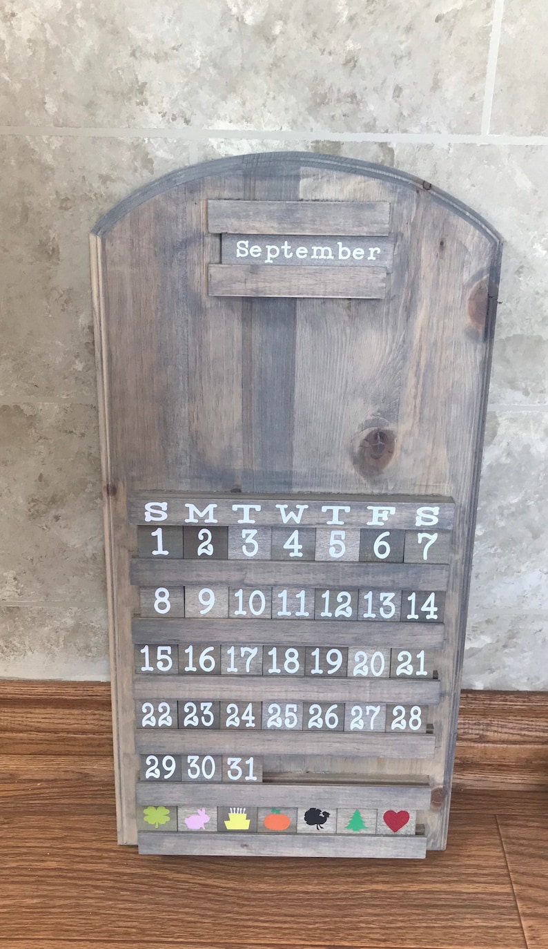 May include: A wooden calendar with a gray wash finish. The calendar features a white inscription for the month of September and a sliding block system for the days of the month. The calendar has a decorative row of icons at the bottom, including a shamrock, a pig, a birthday cake, a pumpkin, a turkey, a Christmas tree, and a heart.