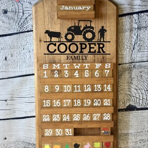 May include: A wooden calendar with a farmhouse theme. The calendar features a tractor, a cow, and a farmer. The calendar is personalized with the name "COOPER FAMILY" and the month of January. The calendar has a slot for each day of the month and includes a small wooden block for each day. The calendar also has a small wooden block for the 4th of July.