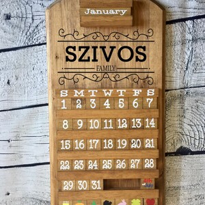 May include: A wooden calendar with the word "January" at the top, the name "SZIVOS FAMILY" in the center, and the days of the week and numbers 1 through 31 below. The calendar has a small wooden shelf at the bottom with colorful wooden blocks.