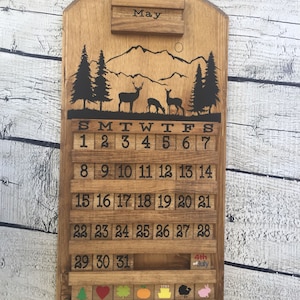 May include: Wooden calendar with a mountain scene and deer silhouette. The calendar is for the month of May and has a small slot for a note. The calendar has a 4th of July decoration.