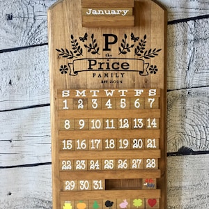 May include: A wooden calendar with a white background and black lettering. The calendar is decorated with a floral design and the text "P the Price Family Est 2014". The calendar has a slot for each day of the month and a small slot for the month name. The calendar is decorated with a small American flag and the text "4th of July".