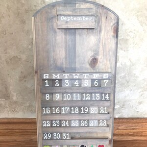 May include: A wooden calendar with a gray wash finish. The calendar features a white inscription for the month of September and white numbers for each day of the month. The calendar has a small shelf with colorful decorations.