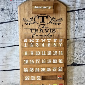 May include: A wooden calendar with the word "January" at the top. The calendar is decorated with a wreath of leaves and the text "the TRAVIS family". The calendar has a letter "T" in a circle at the top. The calendar has a slot for each day of the month and a small slot at the bottom for a special occasion.