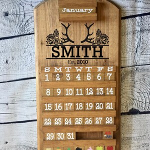 May include: Wooden calendar with a rustic brown finish. The calendar features a personalized name "SMITH" with the year "Est. 2010" and a design of antlers and floral accents. The calendar has a sliding block system for each day of the month, with a separate block for the month of January. The calendar also includes a small block for the 4th of July.
