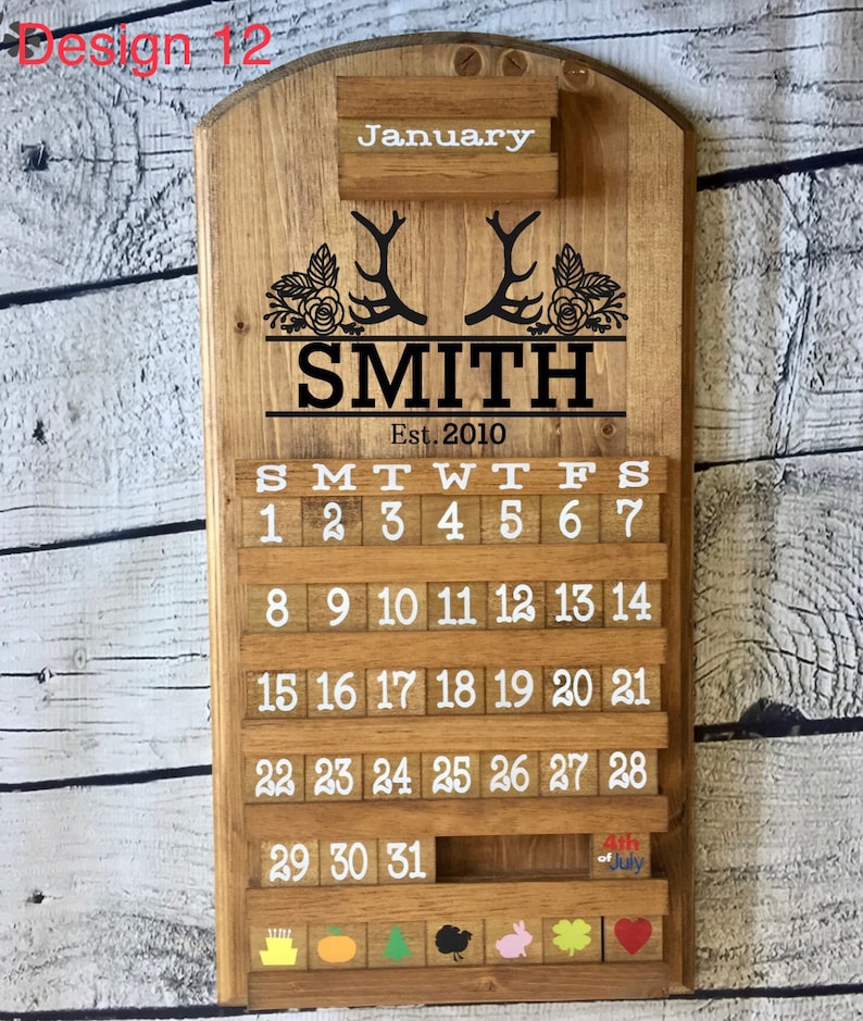 May include: Wooden calendar with a rustic brown finish. The calendar features a personalized name "SMITH" with the year "Est. 2010" and a design of antlers and floral accents. The calendar has a sliding block system for each day of the month, with a separate block for the month of January. The calendar also includes a small block for the 4th of July.