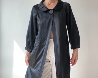 Trench coat longo em cetim chique de seda e algodão com gola Peter Pan. Jaqueta trench coat preta com painel minimalista.