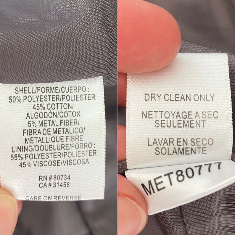 Pode incluir: Close-up de duas etiquetas brancas de vestu&aacute;rio com texto em v&aacute;rios idiomas. As etiquetas detalham a composi&ccedil;&atilde;o do tecido, incluindo poli&eacute;ster, algod&atilde;o e fibra met&aacute;lica. Uma etiqueta indica "Limpar a seco somente".