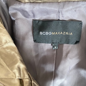 Pode incluir: Close-up de um casaco cor de bronze com forro cinza. A etiqueta preta diz "BCBGMAXAZRIA" em letras douradas. A etiqueta de tamanho diz "S".
