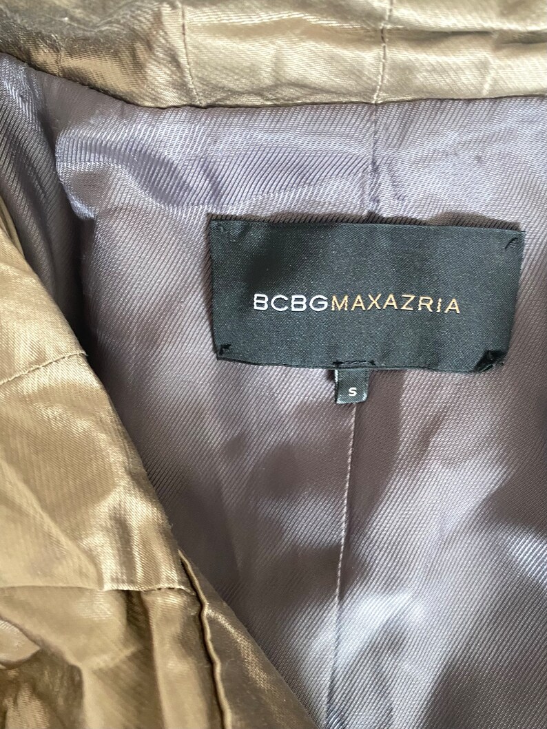 Pode incluir: Close-up de um casaco cor de bronze com forro cinza. A etiqueta preta diz "BCBGMAXAZRIA" em letras douradas. A etiqueta de tamanho diz "S".