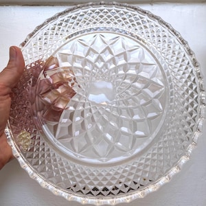 Peut inclure: Une assiette en verre transparent présentant un motif pressé complexe avec un design central en étoile entouré d'anneaux concentriques de facettes en forme de losange. Le bord extérieur est festonné avec un motif en grille de losanges.