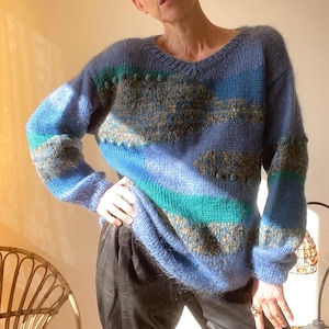 Könnte beinhalten: Ein blau-grün-goldfarbener Strickpullover mit V-Ausschnitt. Der Pullover hat horizontale Streifen und strukturierte Details. Die Ärmel sind lang und der Gesamtstil ist lässig und bequem.