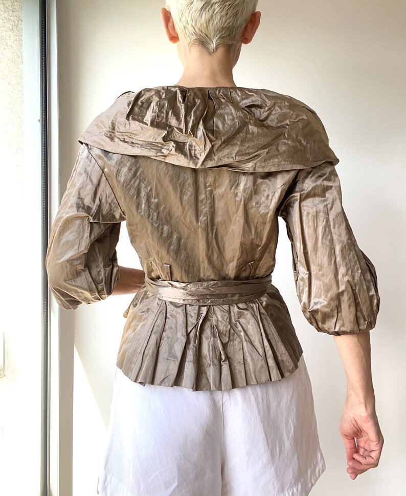 Pode incluir: Top bege de manga curta com gola larga e peplum plissado. O top apresenta um cinto a condizer e &eacute; feito de um tecido brilhante e texturizado. A modelo usa cal&ccedil;&otilde;es brancos.