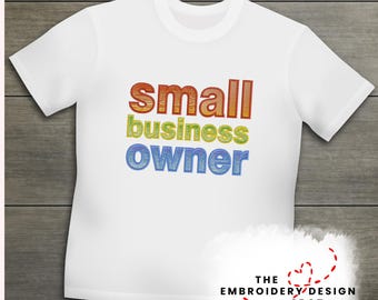 May include: White t-shirt with the text "small business owner" in a rainbow gradient. The text is embroidered on the front of the shirt. The shirt is displayed on a wooden surface.