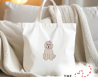 May include: White canvas tote bag featuring an embroidered beige dog with a pink bow. The dog is sitting with its tongue out. The tote bag has two handles and the text "THE EMBROIDERY DESIGN SPOT" at the bottom.