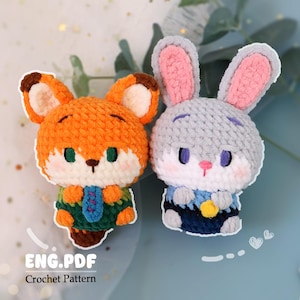 以下が含まれることがあります： オレンジ色のキツネと灰色のウサギの2つの編みぐるみの動物フィギュア。キツネは茶色の耳と緑と青の衣装を着ています。ウサギはピンクの裏地付きの耳と青と黄色の衣装を着ています。「ENG.PDF Crochet Pattern」の文字が見えます。