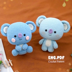 May include: Two light blue crochet koala bears with white and pink accents. The koalas have black eyes and purple noses. The text "ENG.PDF Crochet Pattern" is visible in the image. The koalas are sitting on a brown surface.