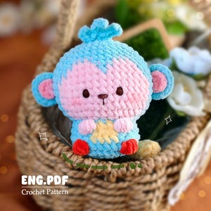 以下が含まれることがあります： 手作りの編み物モンキーおもちゃが柳のバスケットに入っています。モンキーは青とピンクで、お腹に黄色の星、足は赤色です。「ENG.PDF Crochet Pattern」の文字が見えます。
