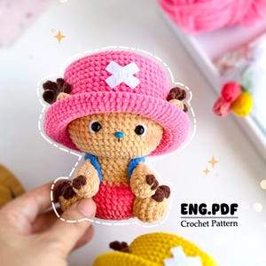 Peut inclure: Une peluche au crochet représentant un personnage de dessin animé, avec un chapeau rose orné d'une croix blanche, des bois bruns et un corps beige. Le jouet porte une tenue rouge avec des bretelles bleues. Le texte "ENG.PDF Crochet Pattern" est visible.