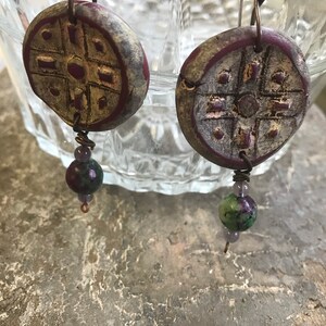 May include: A pair of handmade earrings with a purple and white clay disc with a geometric design and a green and purple stone bead hanging from each disc.